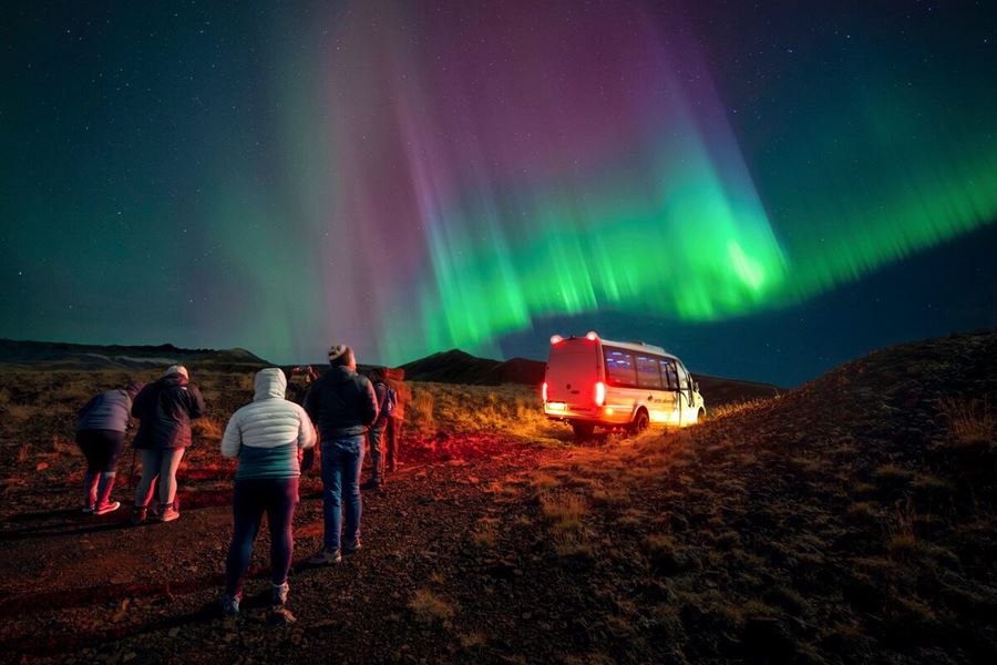 Group of tourists standing next to Arctic Adventures sprinter van underneath northern lights in South West Iceland.