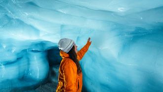 Lady in orange coat reaching to blue ice wall inside ice tunnel 