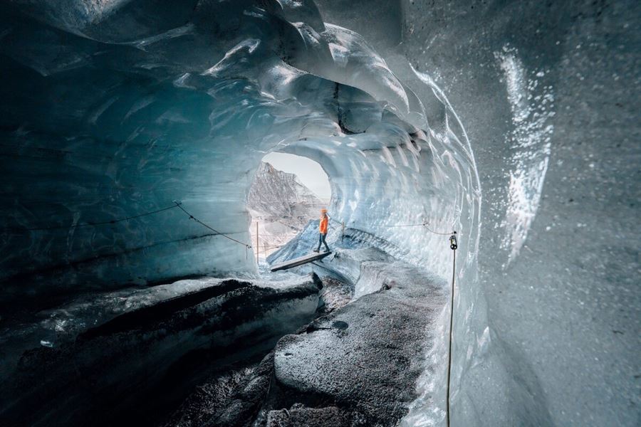 A woman dressed in hiking gear walking past the entrance of the Katla Ice Cave, with blue-tinted ice formations and black volcanic ash layers surrounding the cave opening in Iceland.
