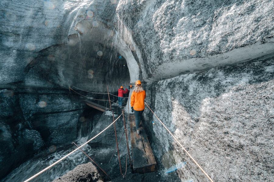 Tourists entering the Katla Ice Cave, stepping through a wide ice tunnel framed by layered volcanic ash and glacial ice in southern Iceland.