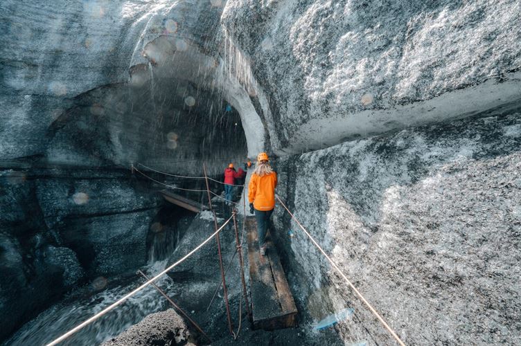 Tourists entering the Katla Ice Cave, stepping through a wide ice tunnel framed by layered volcanic ash and glacial ice in southern Iceland.